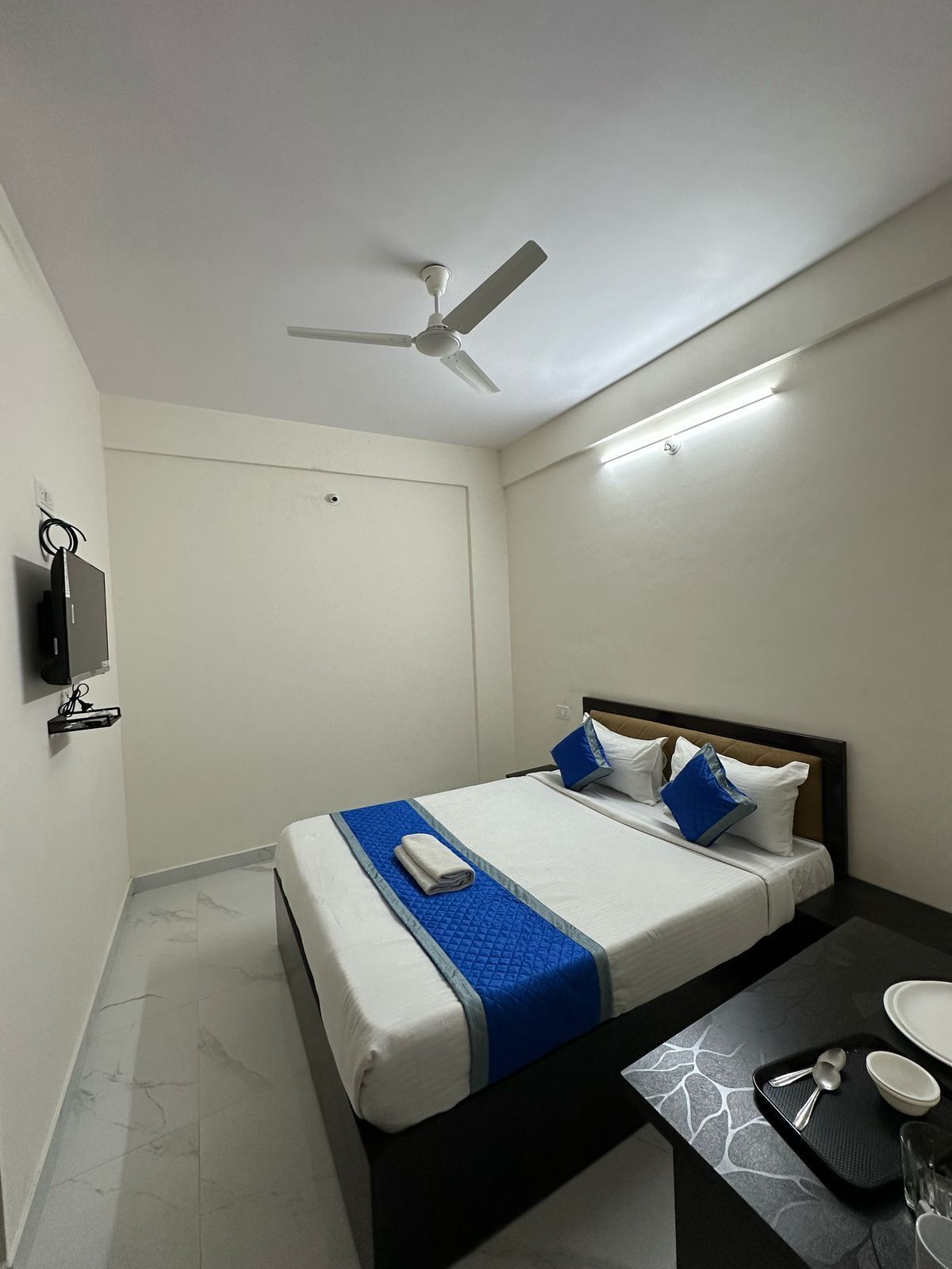 Una habitación de hotel con una cama, una mesita de noche con una bandeja de desayuno, un ventilador de techo y un televisor montado en la pared.