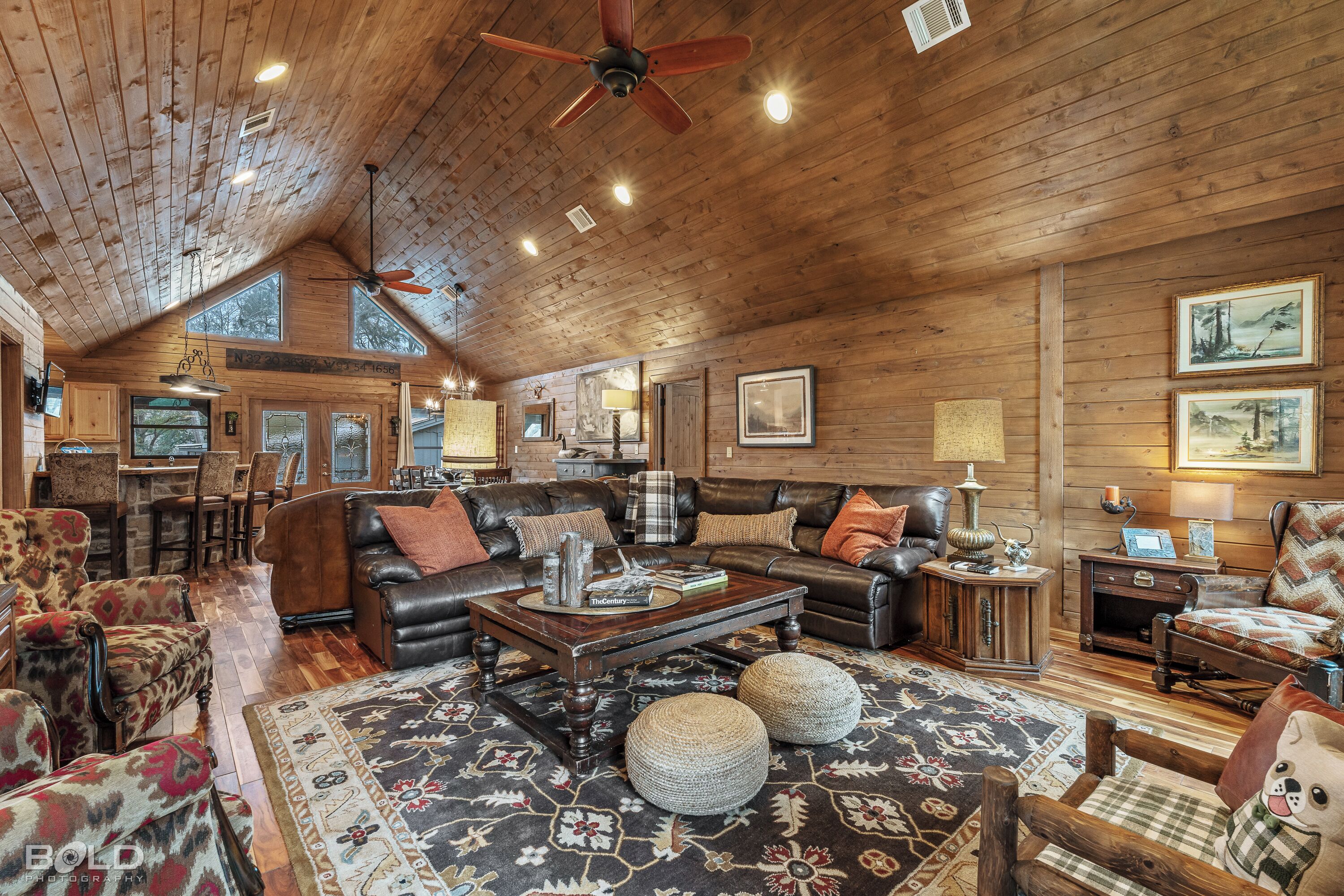 A spacious living room with a high ceiling, wooden walls, and a large window. There are multiple seating areas with leather sofas and armchairs.