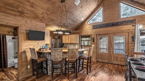 Ein Holzblockhaus-Interieur mit Küchenbereich, einer Bar, Holzbarhocker, einem Flachbildfernseher und einem Esstisch für vier Personen.
