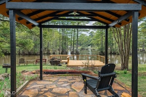A covered outdoor area with a stone patio, a wooden deck, and a view of a lake surrounded by trees. - Relax at this lakefront log cabin on private Emerald Isle, designed for easy outdoor living. Enjoy the hot tub, paddle out with kayaks and canoes, gather around the fire pit, or grill and unwind by the water. (Shreveport)