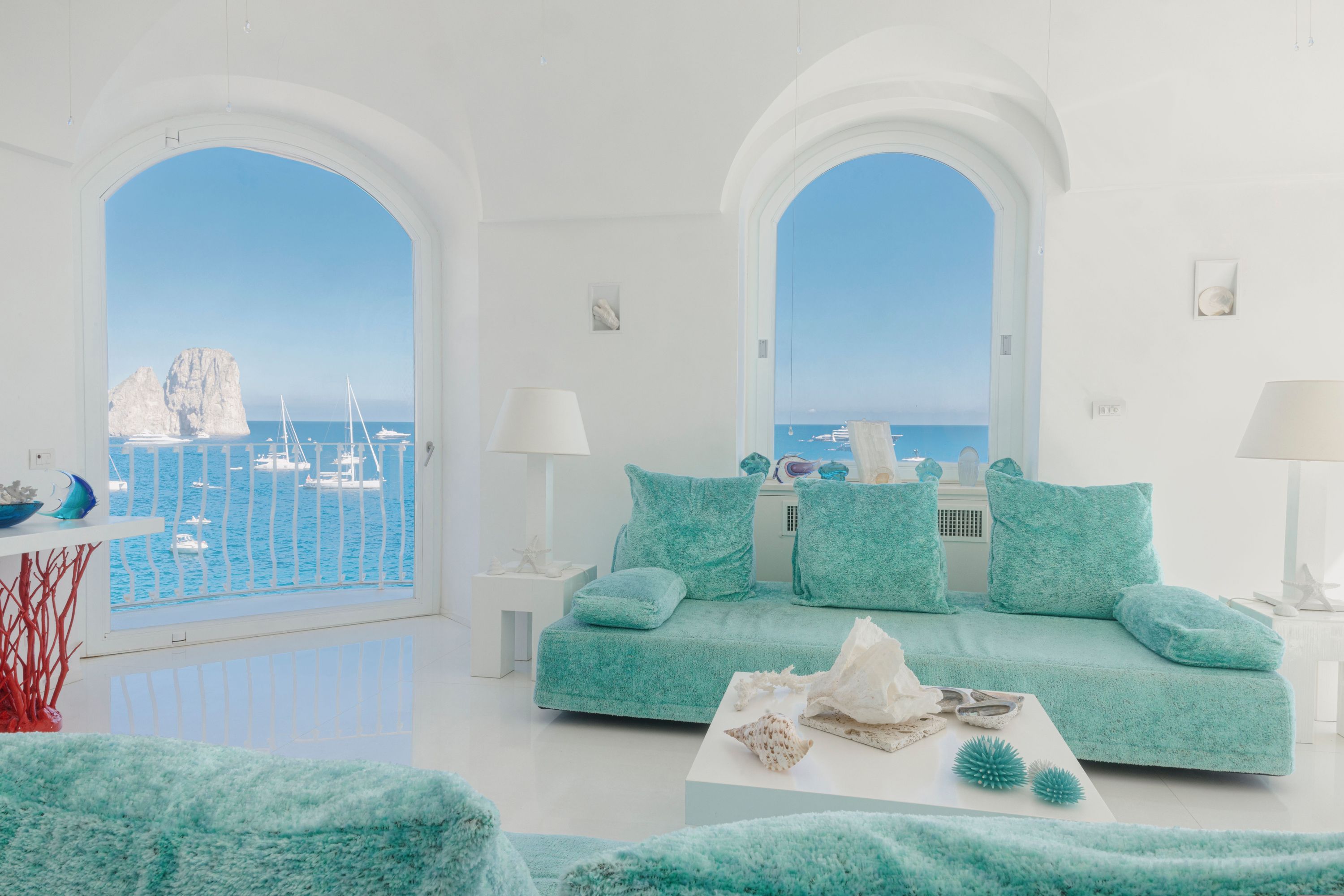 A modern living room with a teal sofa, white lamps, and a view of the sea and boats.