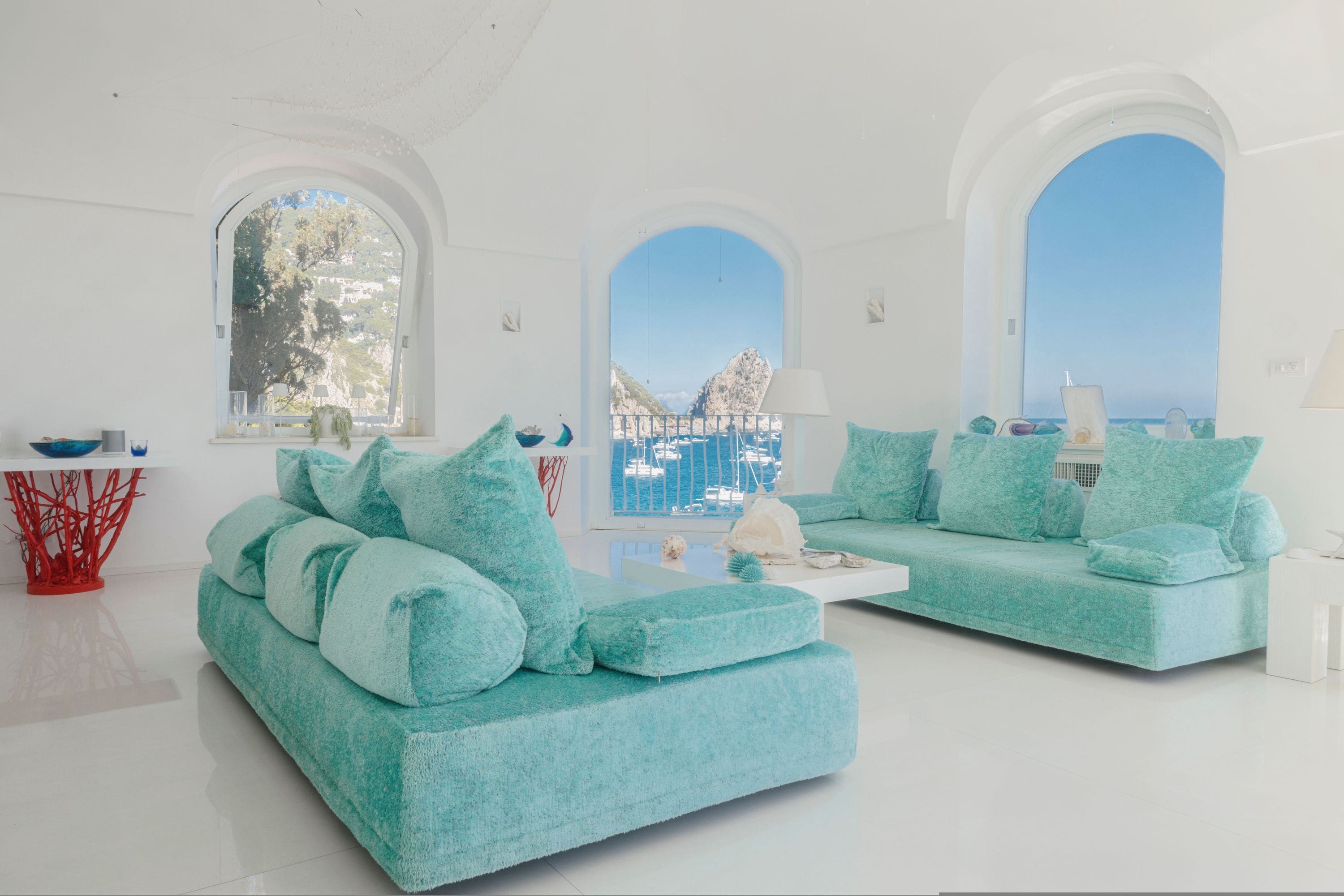 Una sala de estar moderna con dos sofás color turquesa, una mesa de centro blanca y una escultura abstracta roja. Amplias ventanas en arco ofrecen vistas al mar y a las embarcaciones.