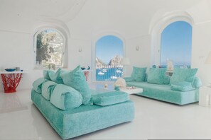 A modern living room with two teal sofas, a white coffee table, and a red abstract sculpture. Large arched windows provide views of the sea and boats. - Villa Beatrice by Elite Villas (Capri)