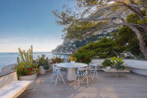 An outdoor patio with a round table and chairs, surrounded by potted plants and overlooking a coastal area with cliffs and the sea. - Villa Beatrice by Elite Villas (Capri)