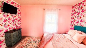 Une chambre avec un papier peint à motifs floraux, un lit avec une literie rose, une commode et un téléviseur fixé au mur.