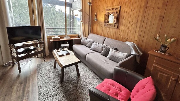 Ein gemütliches Wohnzimmer mit Holzbalkendecke, einem grauen Sofa, einem Couchtisch, einem Fernseher auf einem Holzständer und einem Fenster mit Blick auf Bäume.