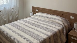 A bedroom with a sloped ceiling, wooden beams, a bed with a striped blanket, two framed pictures on the wall, and a window with a curtain. - Casa Bice - A new apartment in the center of Tesino (Castello Tesino)