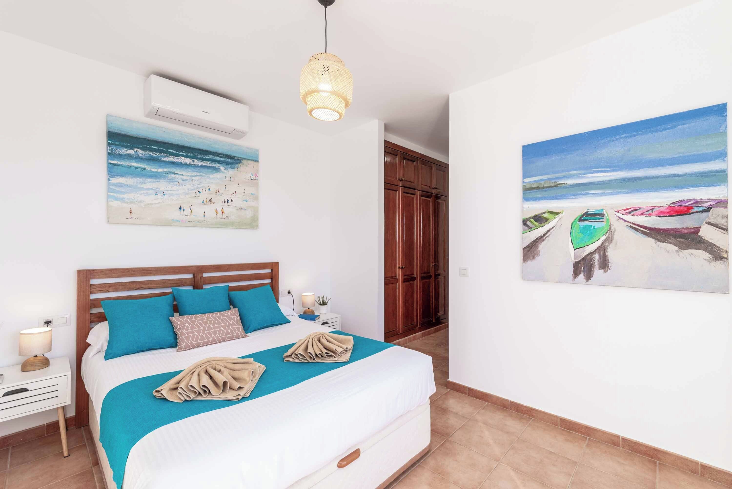 A bedroom with a bed, a nightstand, a wall-mounted air conditioner, a hanging light fixture, and two paintings.