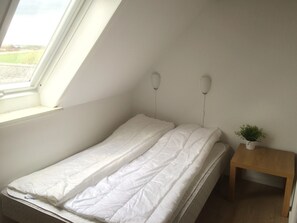 Ein kleines, ordentlich bezogenes Bett in einem Raum mit schrägem Dach und einem Dachfenster. Ein kleiner Nachttisch aus Holz mit einer Pflanze darauf.