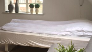 Habitación sencilla y luminosa con cama, ventana y dos plantas en macetas, además de una lámpara de pared.