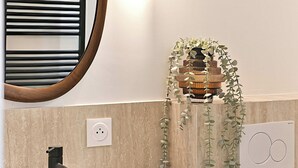 Ein Badezimmer mit weißem Waschbecken, einem schwarzen Wasserhahn, einem Holz-Waschtisch, einem Spiegel mit Holzrahmen, einer wandmontierten Leuchte und einer hängenden Pflanze.