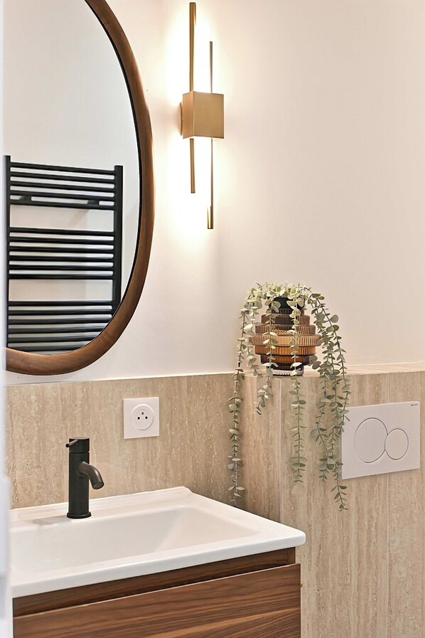 Een badkamer met een witte wastafel, een zwarte kraan, een houten wastafelmeubel, een spiegel met houten lijst, een wandlamp en een hangende plant.