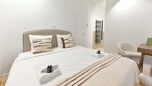 Ein ordentlich bezogenes Bett mit weißen Bettwäsche und einer beige farbenen Decke, ein weißer Stuhl und ein kleiner Holztisch mit einer grünen Lampe.