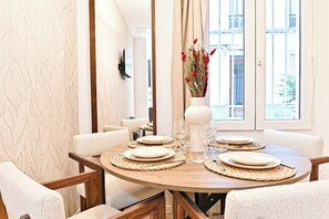 Een eetruimte met een ronde tafel voor vier personen, omringd door witte fauteuils. Op de tafel staat een vaas met rode bloemen, en er is een groot raam met uitzicht op gebouwen.