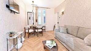 Ein modernes Wohnzimmer mit Fischgrätparkett, einem grauen Sofa, einem Essbereich mit Tisch und Stühlen und einem großen Fenster.