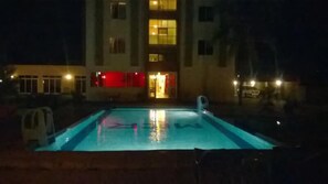 Una piscina nocturna con un edificio al fondo.