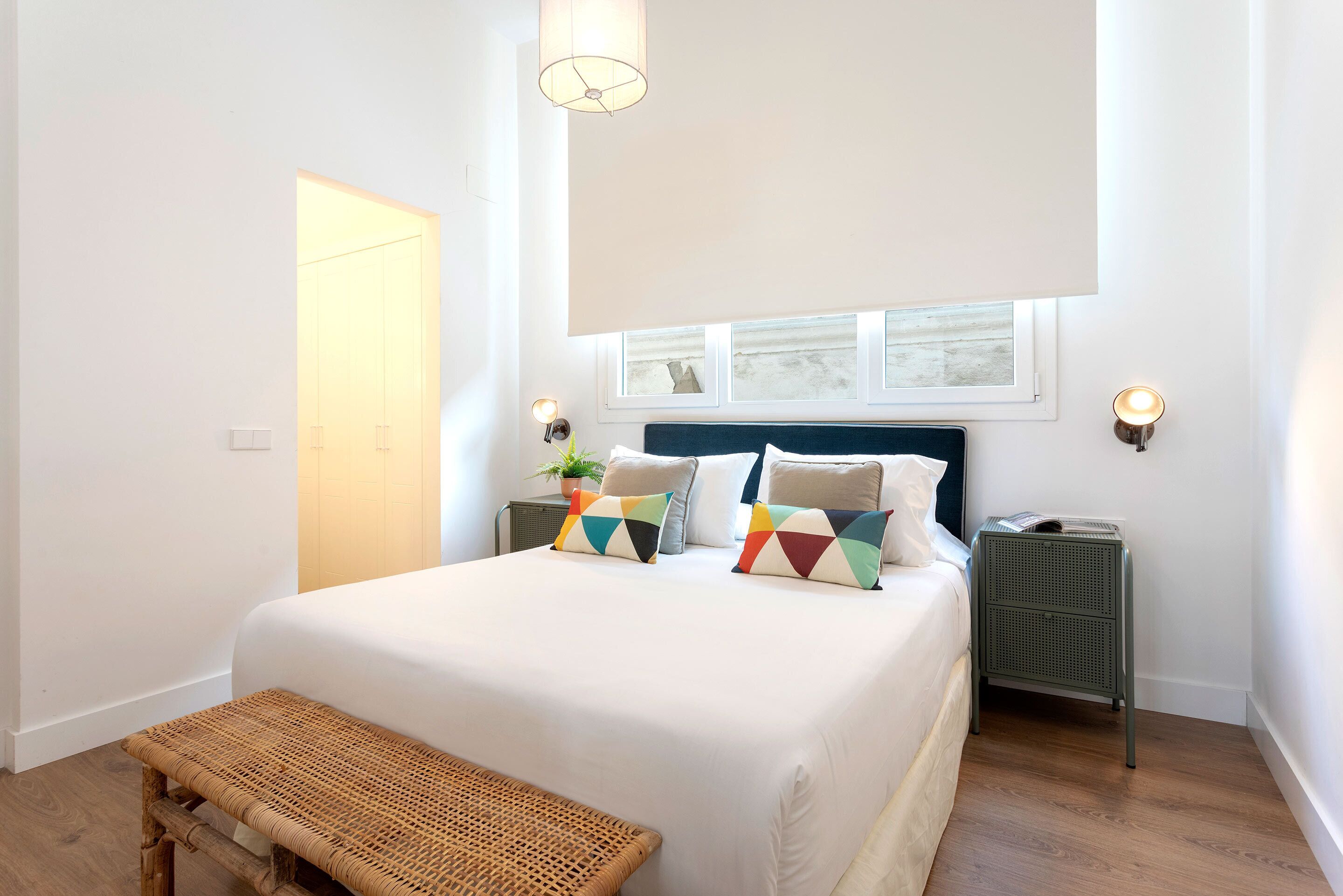 A bedroom with a large bed, two bedside tables, a bench, and a window with blinds.