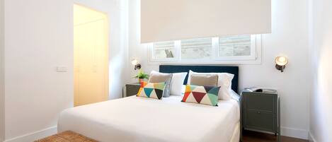 A bedroom with a large bed, two bedside tables, a bench, and a window with blinds.