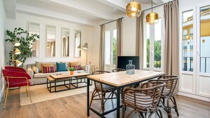 Ein geräumiger Wohnbereich mit einem Holzessttisch, Korbstühlen, einem roten Sessel und einer Couch mit bunten Kissen.