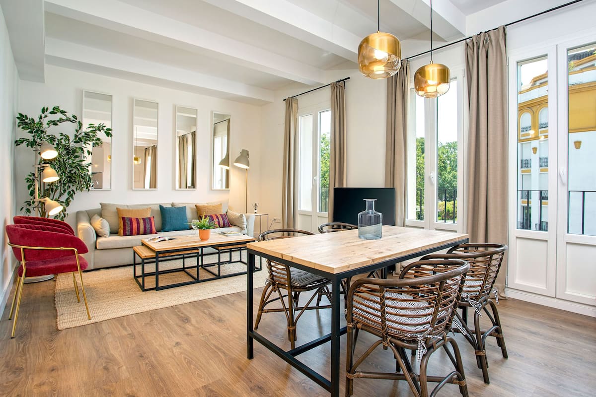A spacious living area with a wooden dining table, wicker chairs, a red armchair, and a sofa with colorful cushions.
