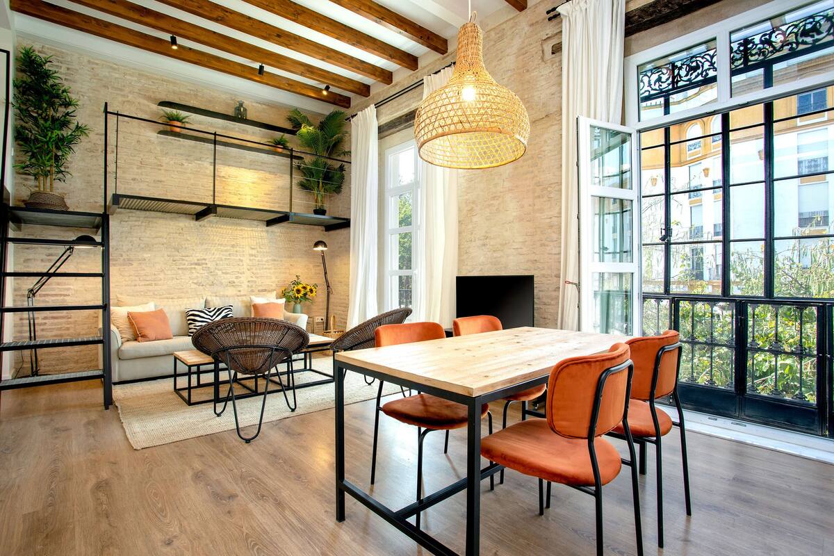 A dining area with a wooden table and orange chairs, a hanging light fixture, and a brick wall.