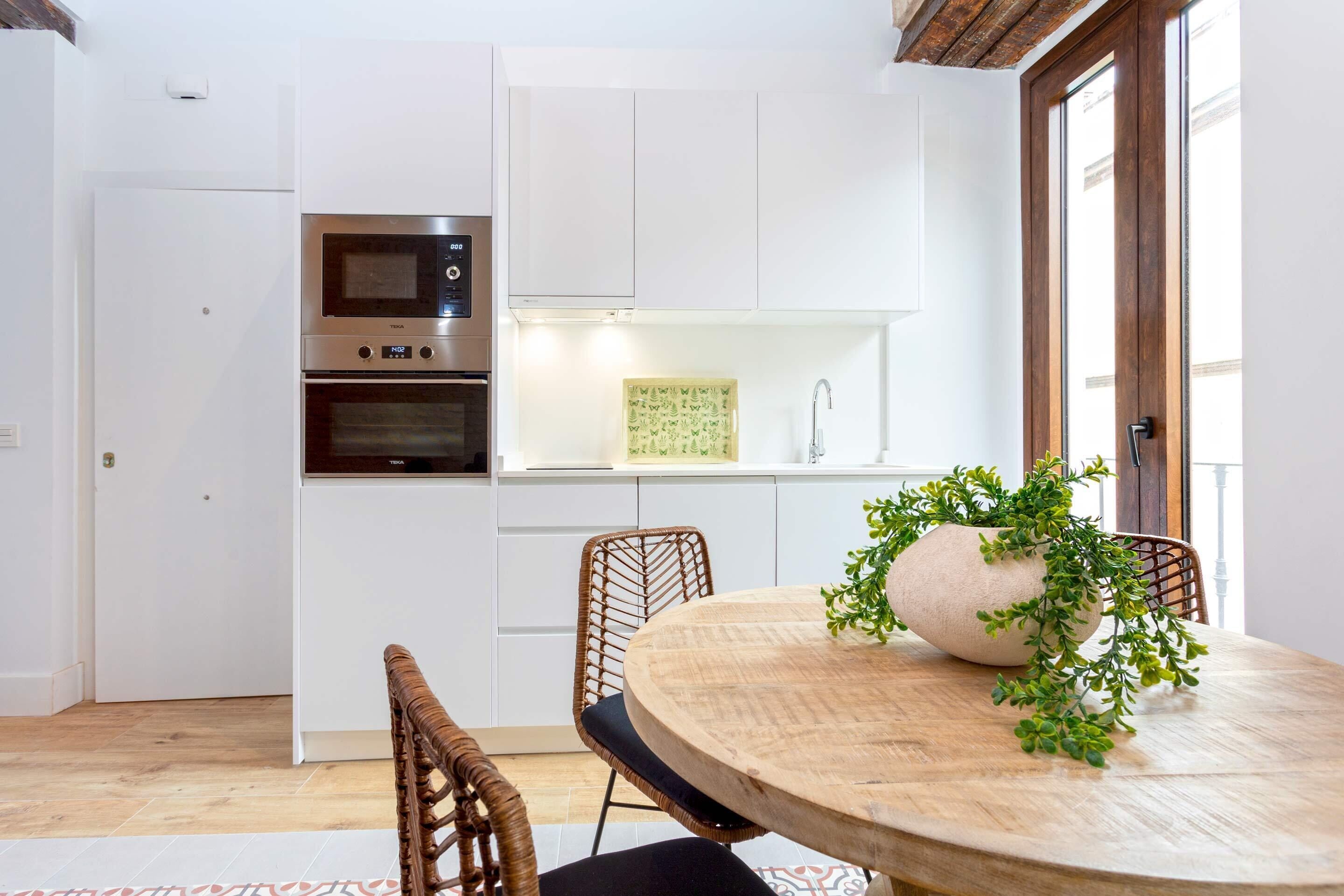 A modern kitchen with white cabinetry, a built-in oven, and a dining area with a round wooden table and wicker chairs.
