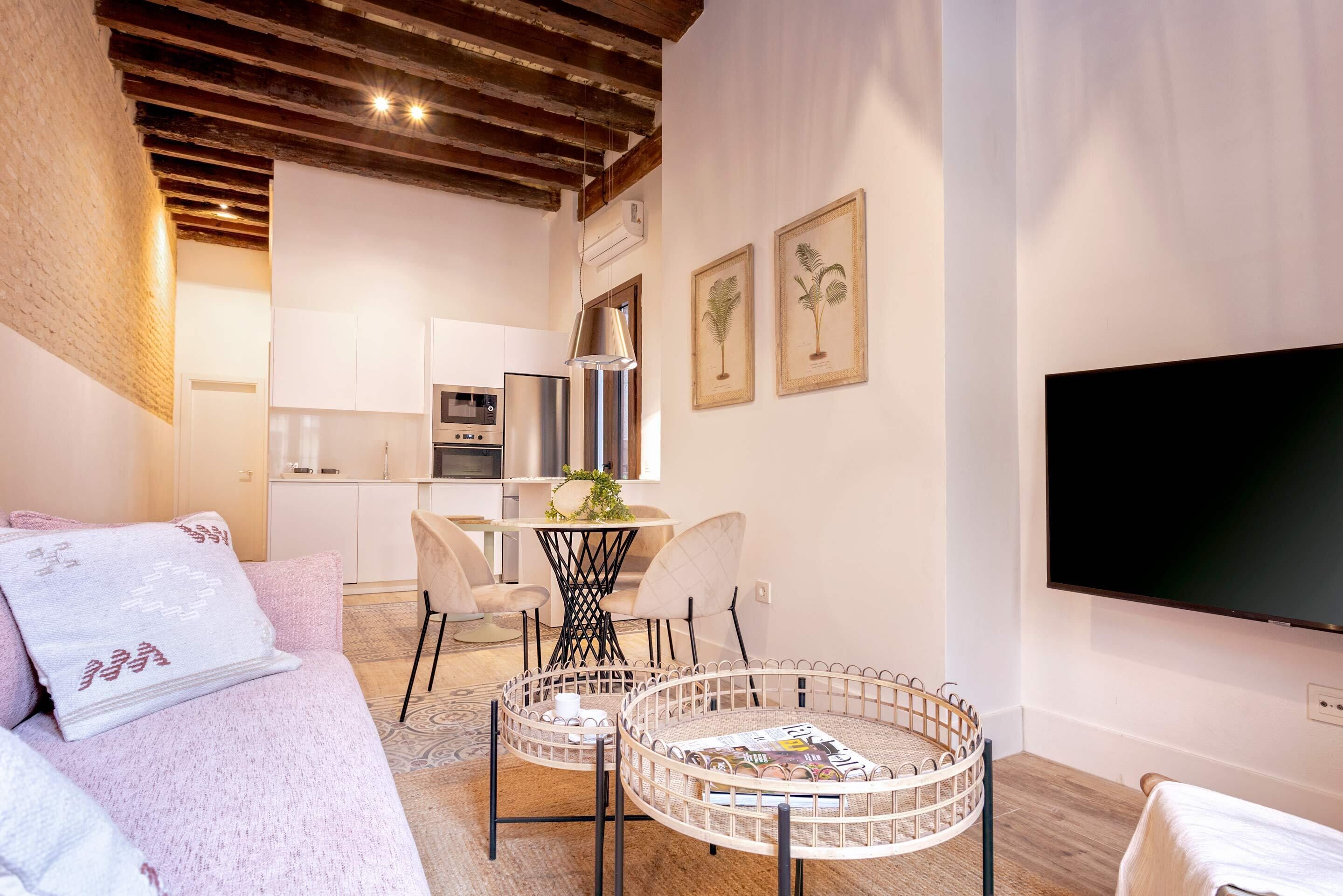 A modern living room with a sofa, a coffee table, and a TV. There is a kitchen in the background with cabinets and appliances.