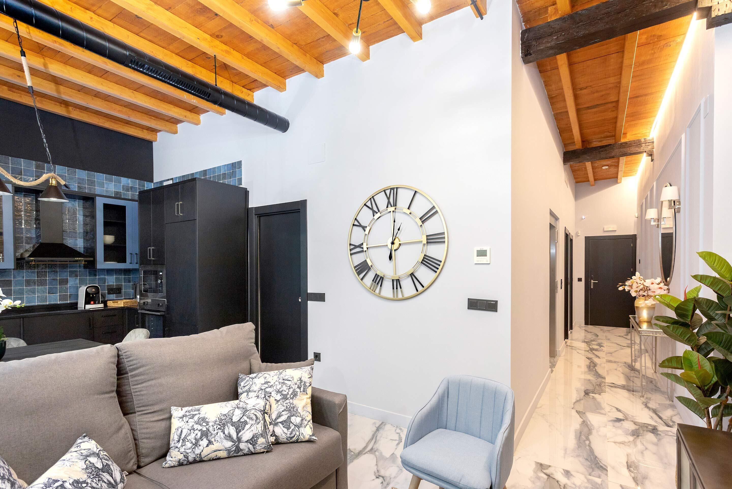 Un intérieur moderne avec un espace de vie comprenant un canapé et un fauteuil bleu, une grande horloge murale, et une cuisine dotée de meubles sombres et de carreaux bleus.