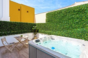 Un bain à remous dans une cour, avec une table et des chaises en bois, entouré d’une haie et d’un mur jaune.