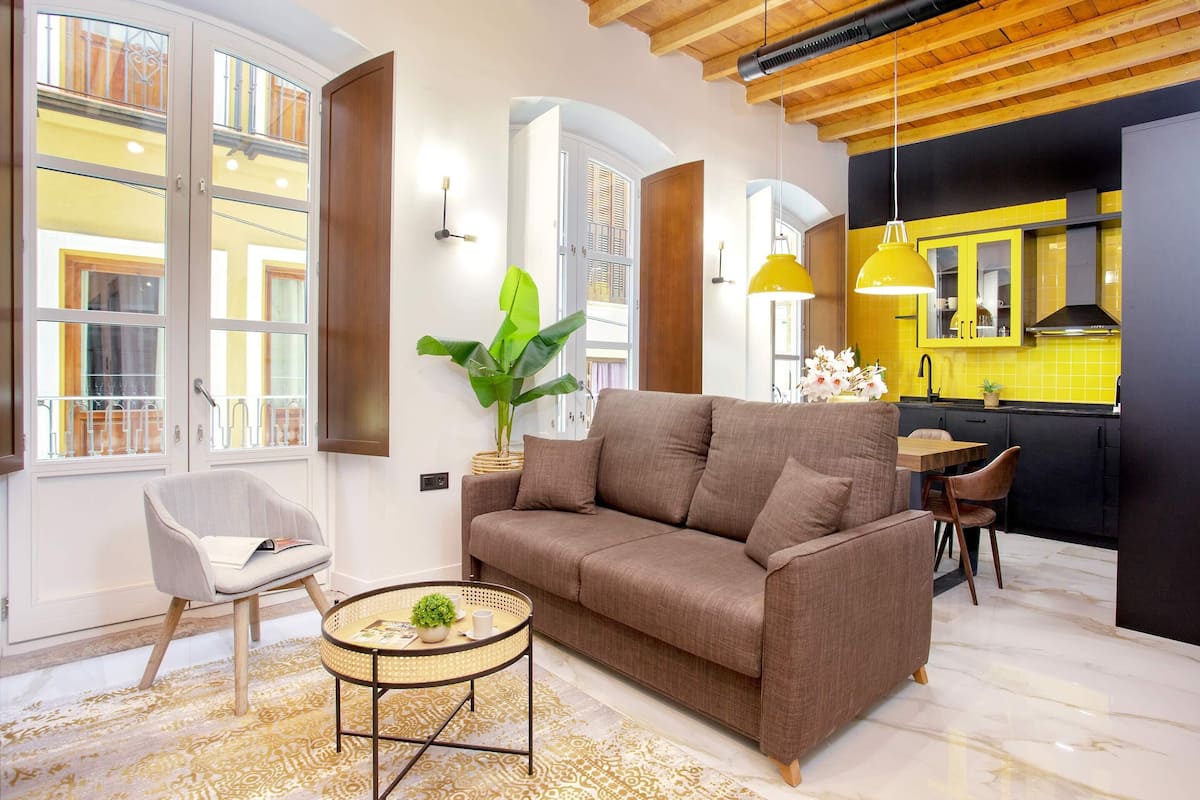 A living room with a sofa, a coffee table, and a chair. There is a kitchen area with yellow cabinets and a sink.