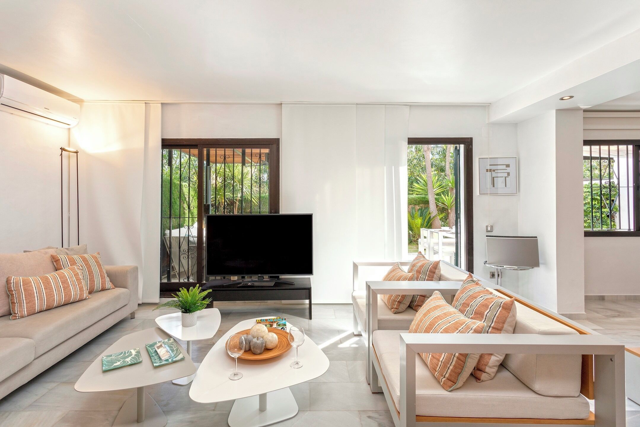 A modern living room with a sofa, a coffee table, a TV, and a balcony with a view of greenery.