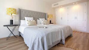 Ein Schlafzimmer mit grau gestepptem Kopfteil, weißer Bettwäsche, zwei Nachttischen mit Lampen und einem großen Kleiderschrank.