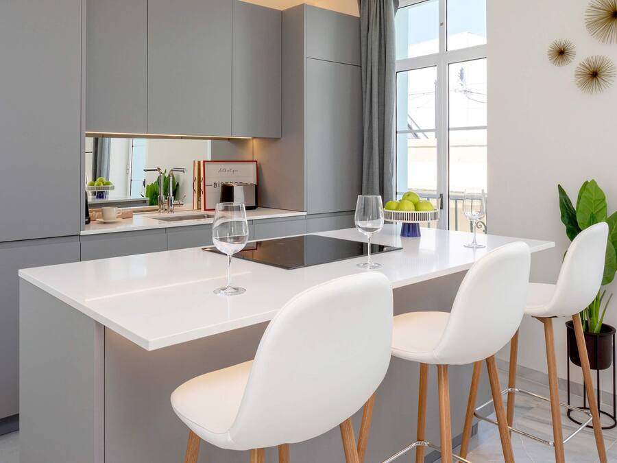 A modern kitchen with a central island, white bar stools, and a large window.