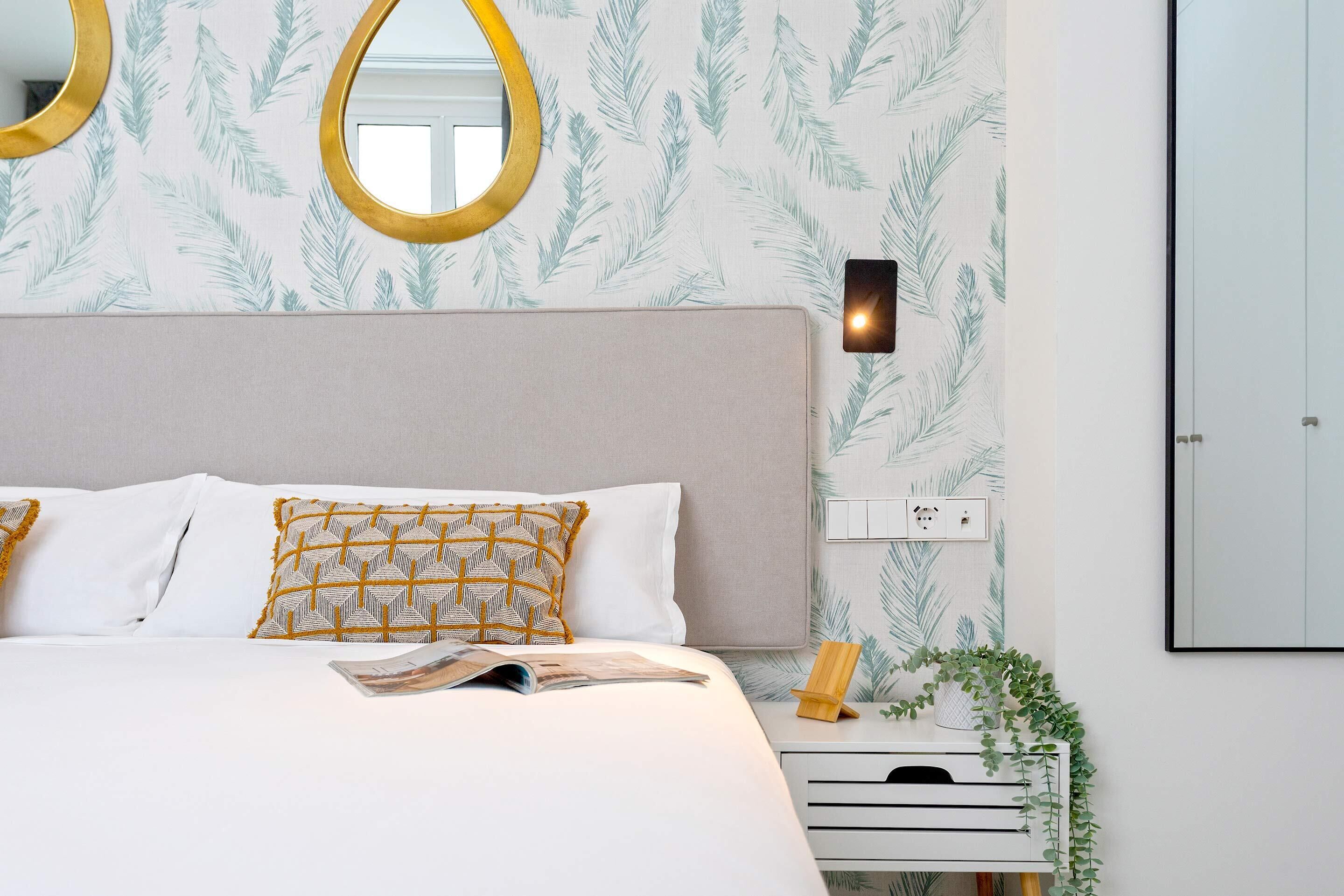 Una cama bien hecha con almohadas blancas y estampadas, una mesita de noche con lámpara y un espejo montado en la pared.