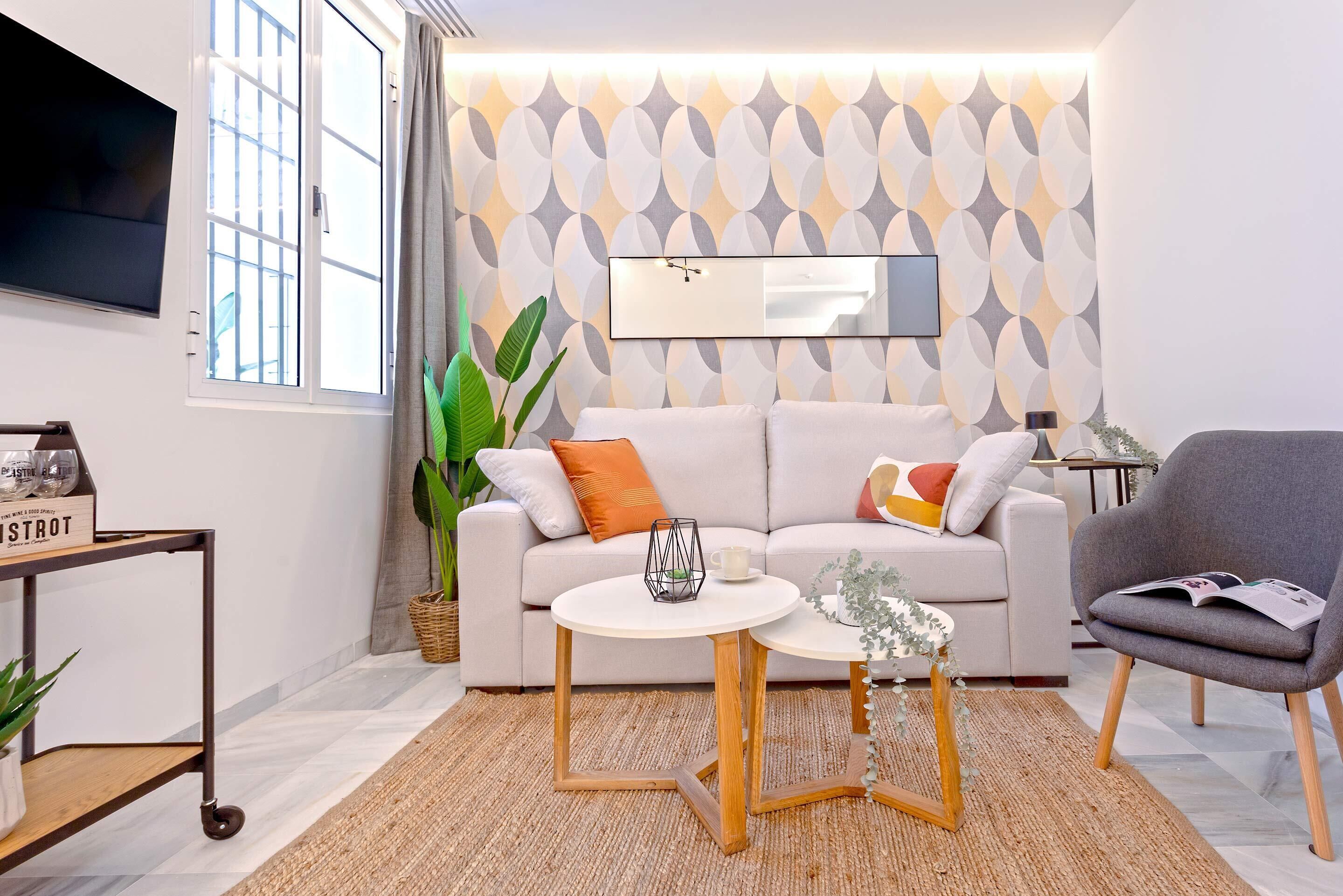 Una sala de estar moderna con un sofá, dos mesitas auxiliares, un sillón gris, un ventanal grande y una pared con diseño.