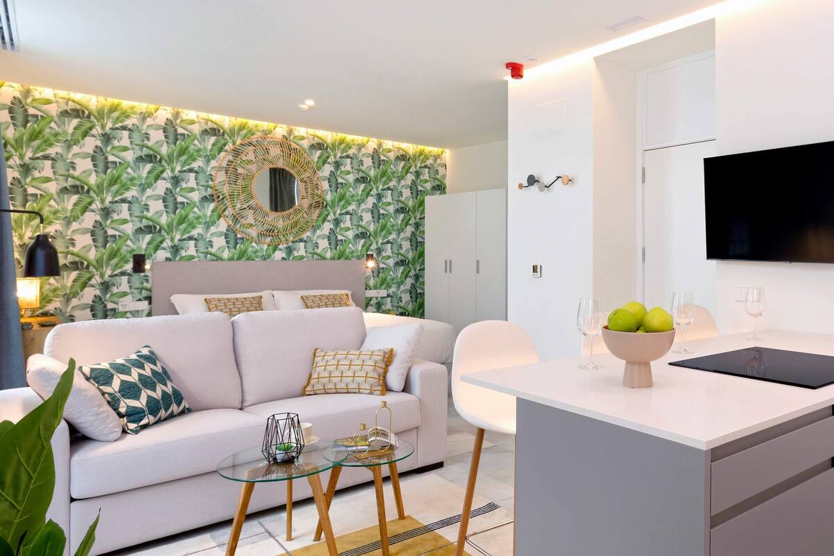 Un salon moderne avec un canapé blanc, une table basse en verre et un coin cuisine doté d’un plan de travail blanc et d’un téléviseur à écran plat.