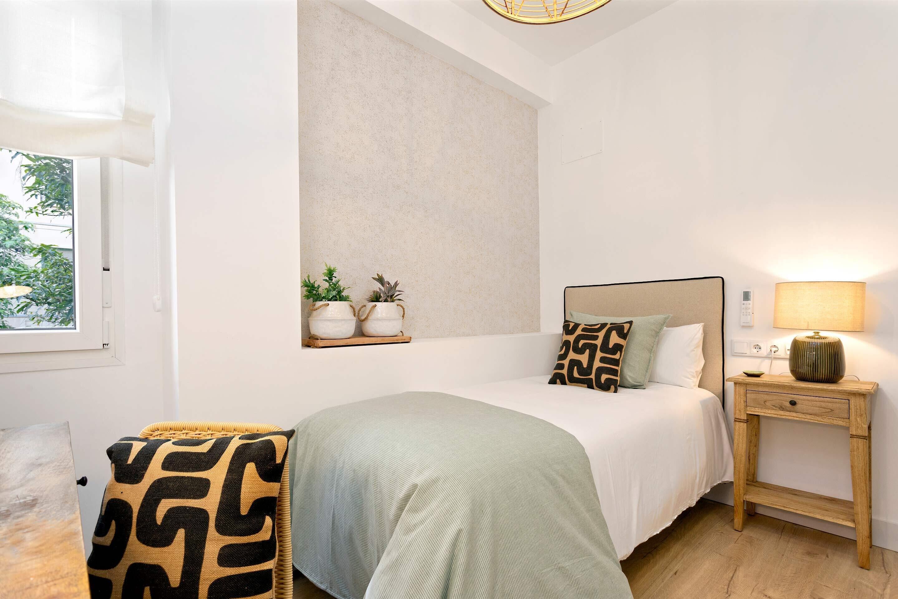 Een slaapkamer met een bed, nachtkastje, lamp en een raam met uitzicht op groen.