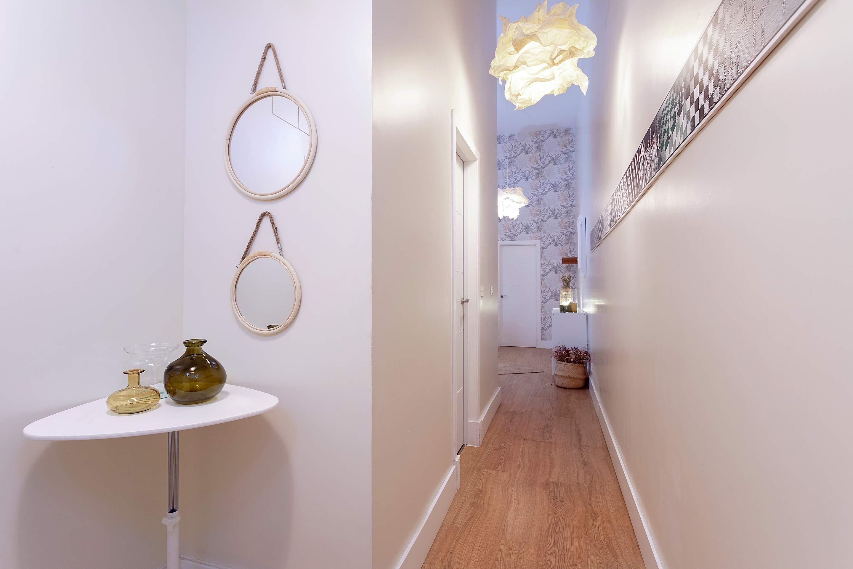 A hallway with a round table, two wall-mounted mirrors, and a hanging light fixture.