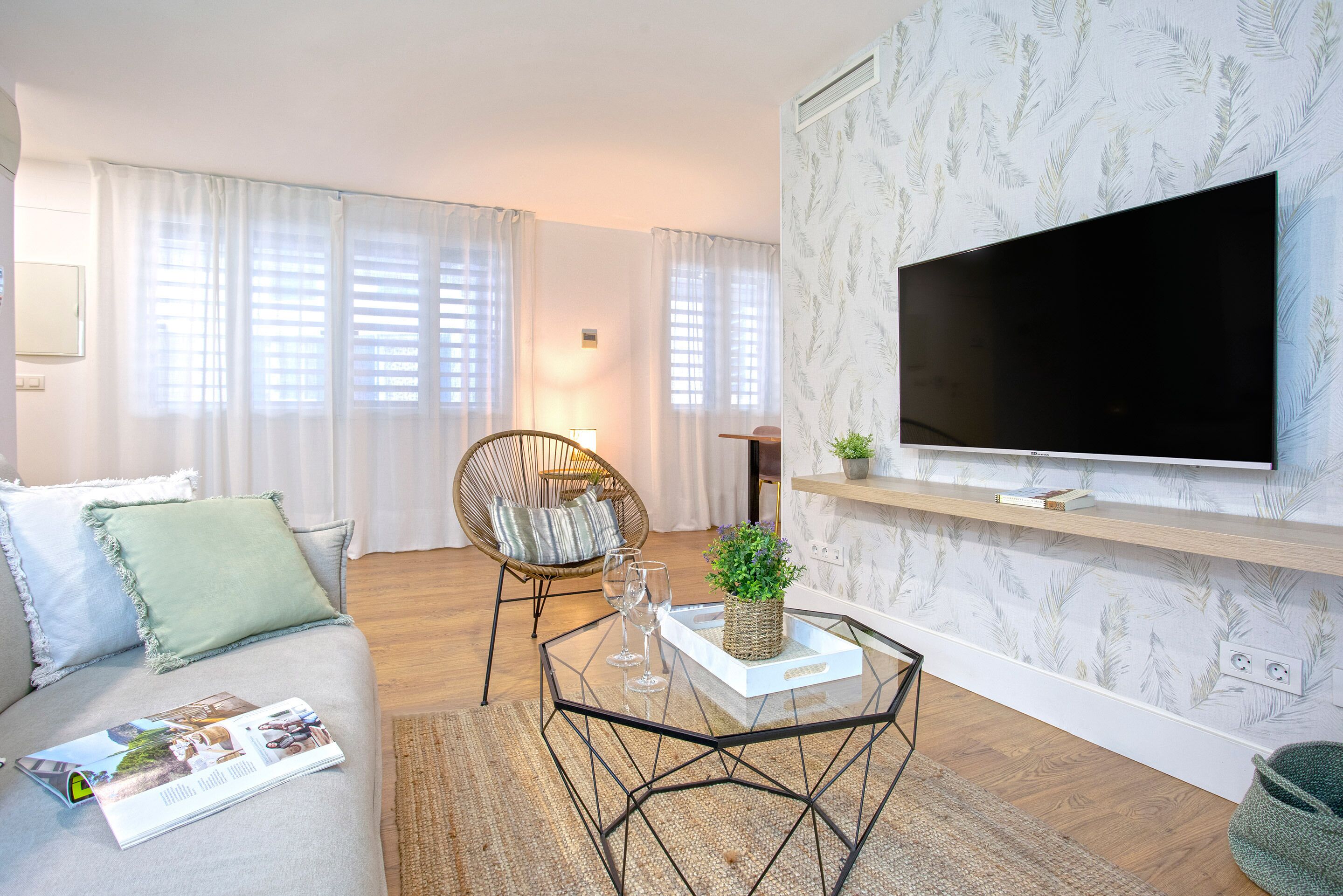 A modern living room with a sofa, a glass coffee table, a TV, and a patterned wallpaper.