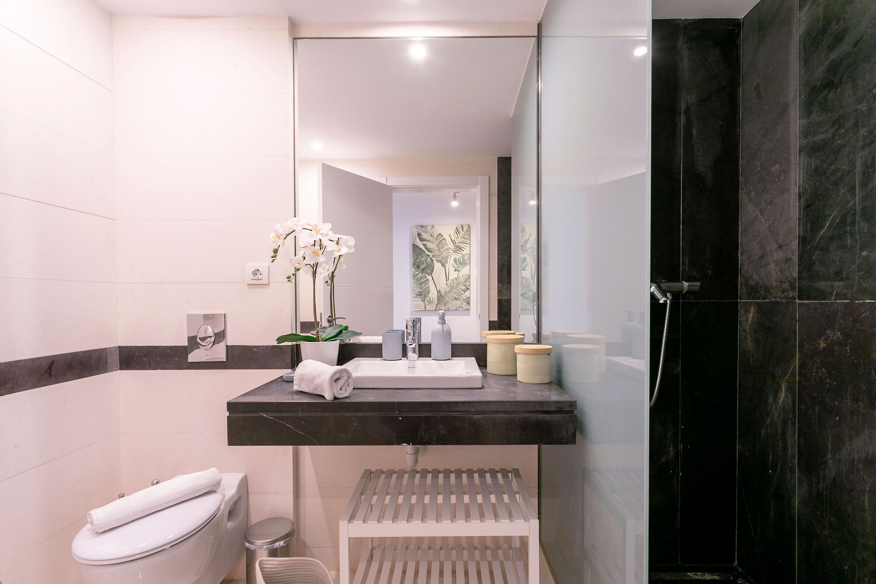 A modern bathroom with a black marble shower, a white sink with a mirror, and a small white shelf with toiletries.