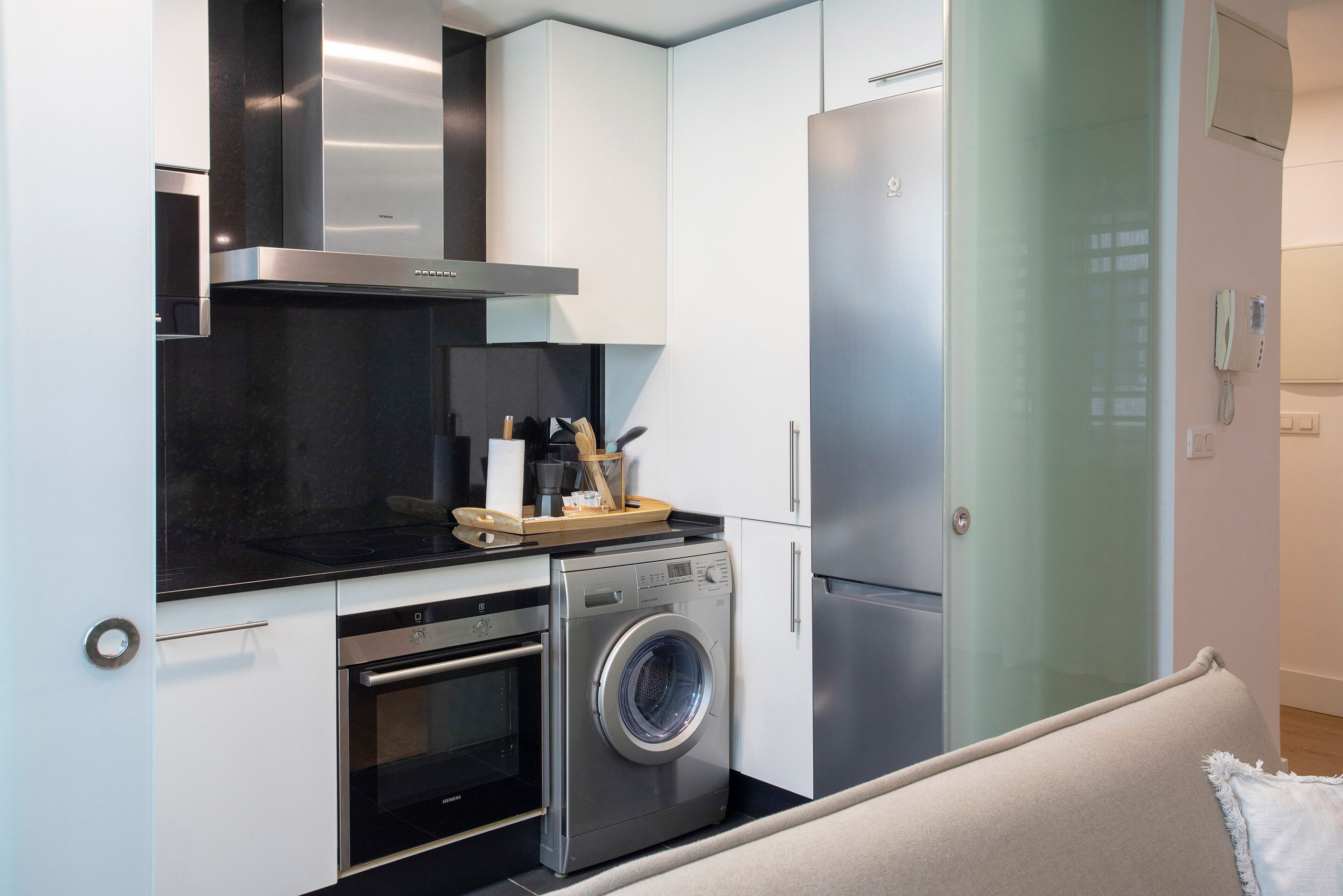 A compact kitchen with a stainless steel refrigerator, a built-in oven, a washing machine, and a coffee machine.