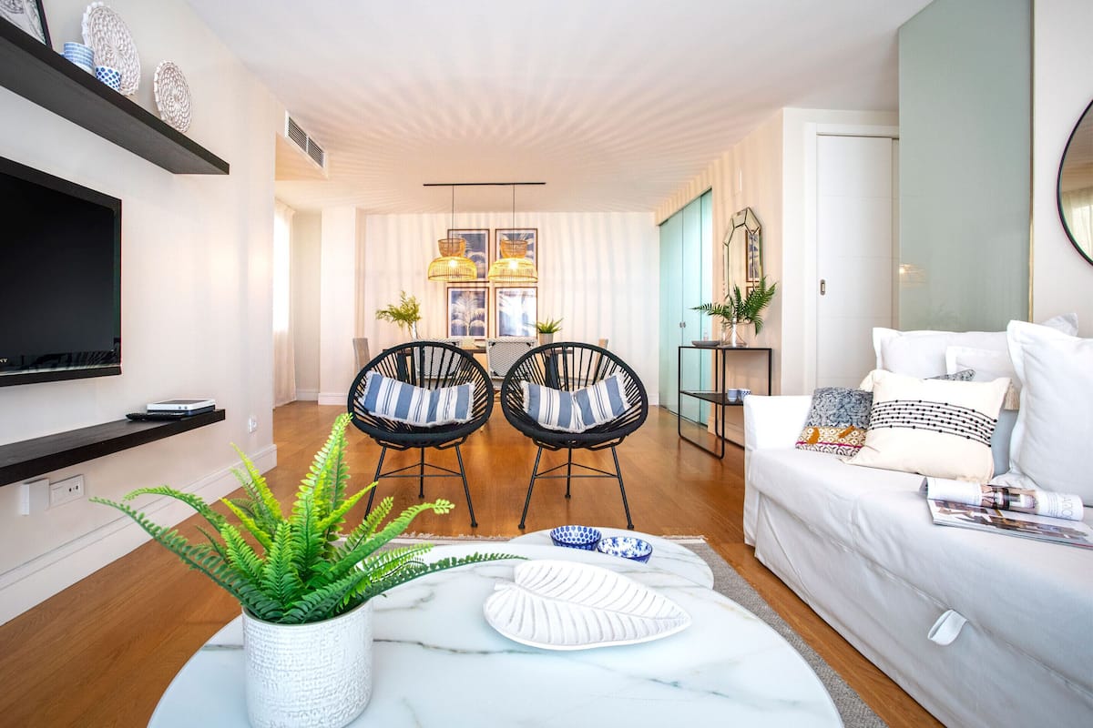 A modern living room with a flat-screen TV, two black chairs with striped cushions, a white sofa with patterned pillows, and a dining area with a table and chairs.