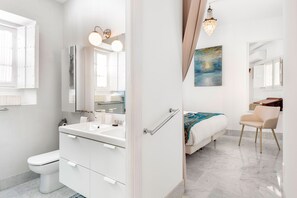 Un baño moderno con lavamanos, inodoro y tocador blancos. En la habitación contigua, hay una cama con manta azul y una silla.