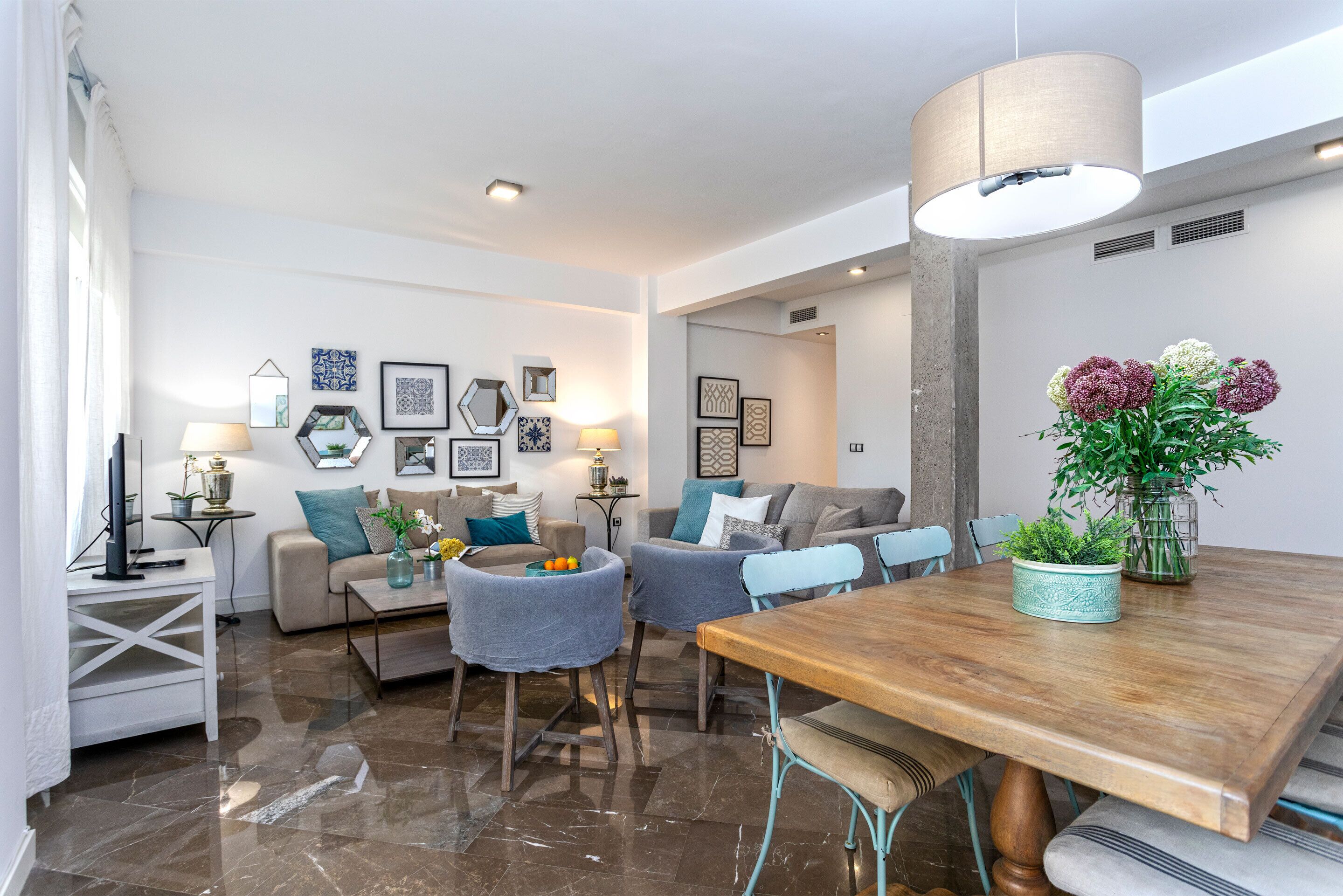 A modern living room with a wooden dining table, grey sofas, and a coffee table. There are framed artworks on the walls and a vase with flowers on the table.