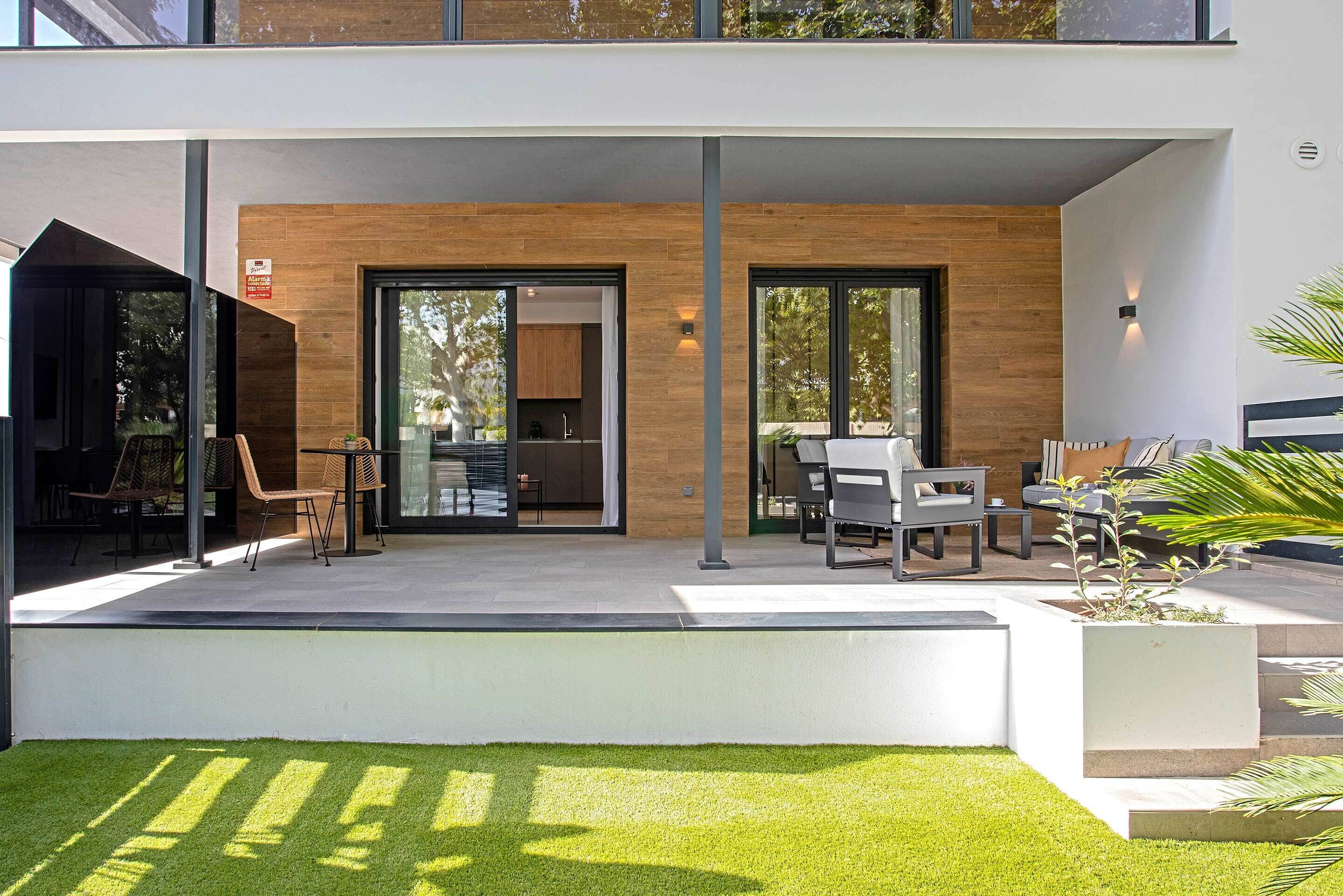 Un patio exterior moderno con zona de comedor, cocina visible a través de puertas de vidrio y un césped bien cuidado.