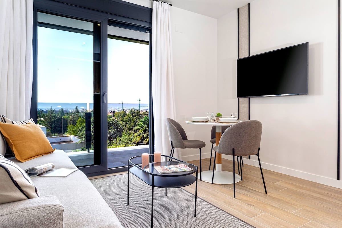 Una sala moderna con un sofá, una mesa de centro y un comedor con sillas. Un televisor de pantalla plana está montado en la pared.