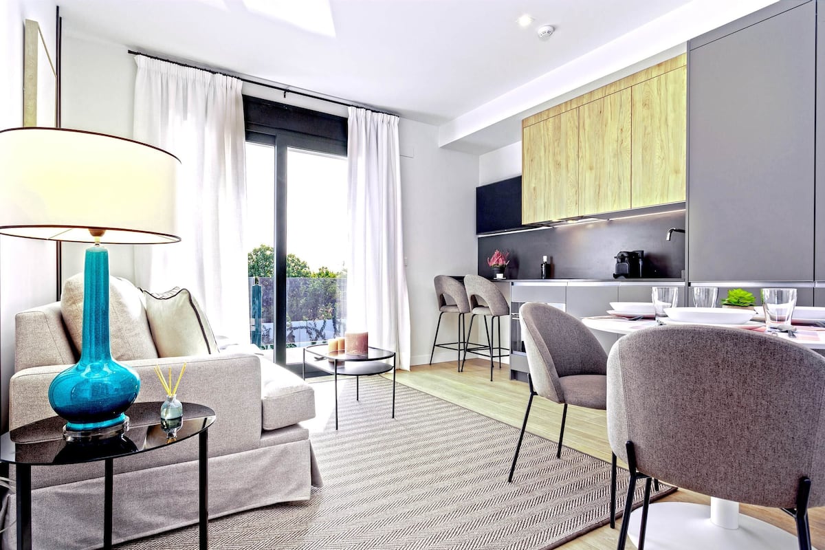 Una sala de estar moderna con sofá, mesa de centro y lámpara. Hay un comedor con mesa y sillas. La habitación tiene un ventanal con cortinas.