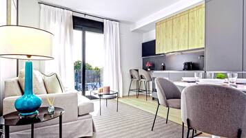 A modern living room with a sofa, coffee table, and lamp. There is a dining area with a table and chairs. The room has a large window with curtains.