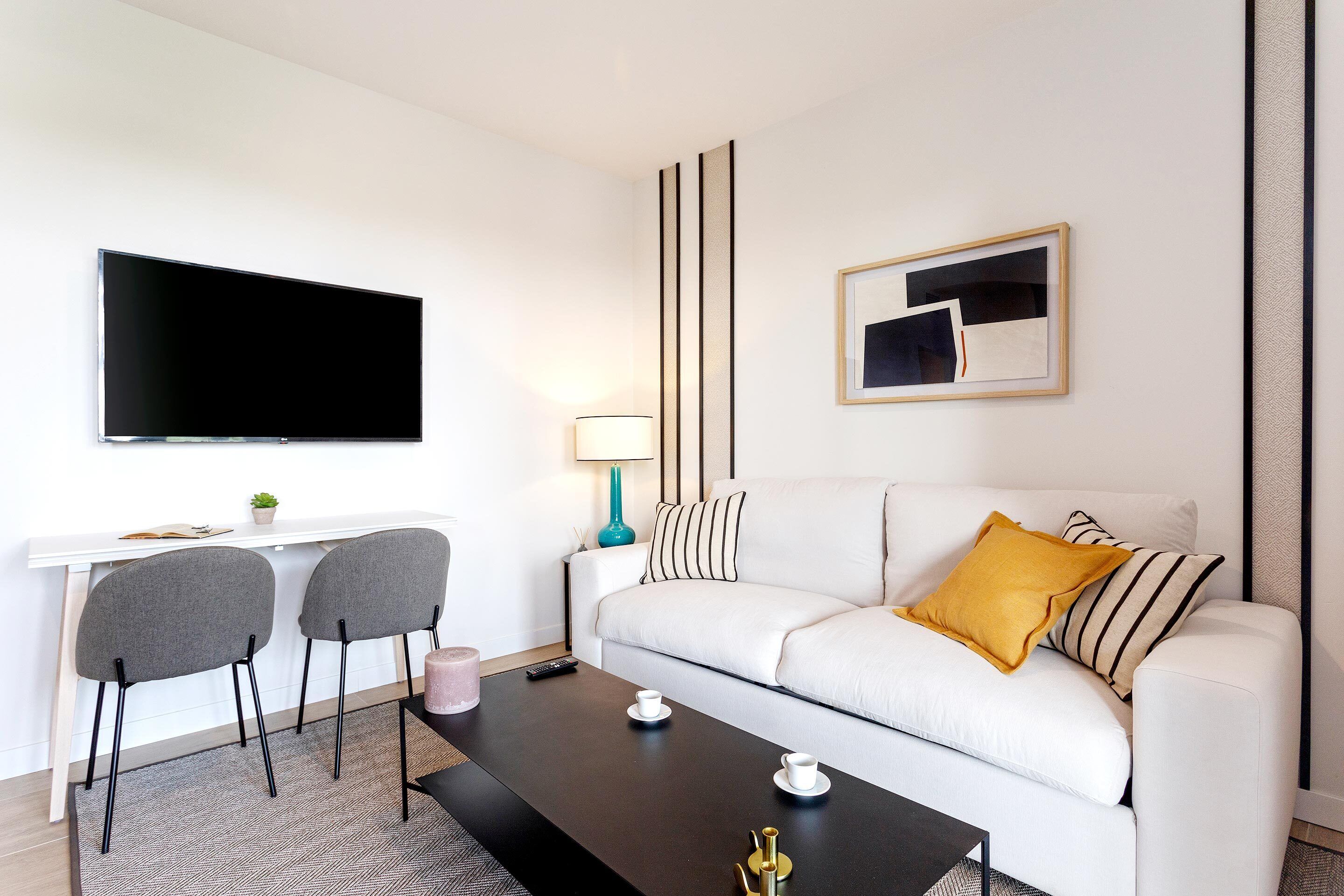 Una sala de estar moderna con un sofá blanco, dos sillones grises, una mesa de centro negra, un televisor de pantalla plana, un escritorio con lámpara y un cuadro abstracto enmarcado.