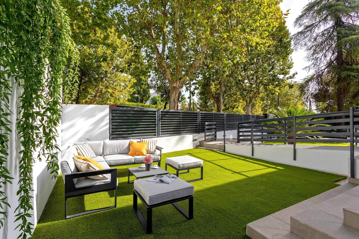 Un área de estar moderna al aire libre con un sofá blanco, dos mesas auxiliares negras y una mesita con un jarrón.